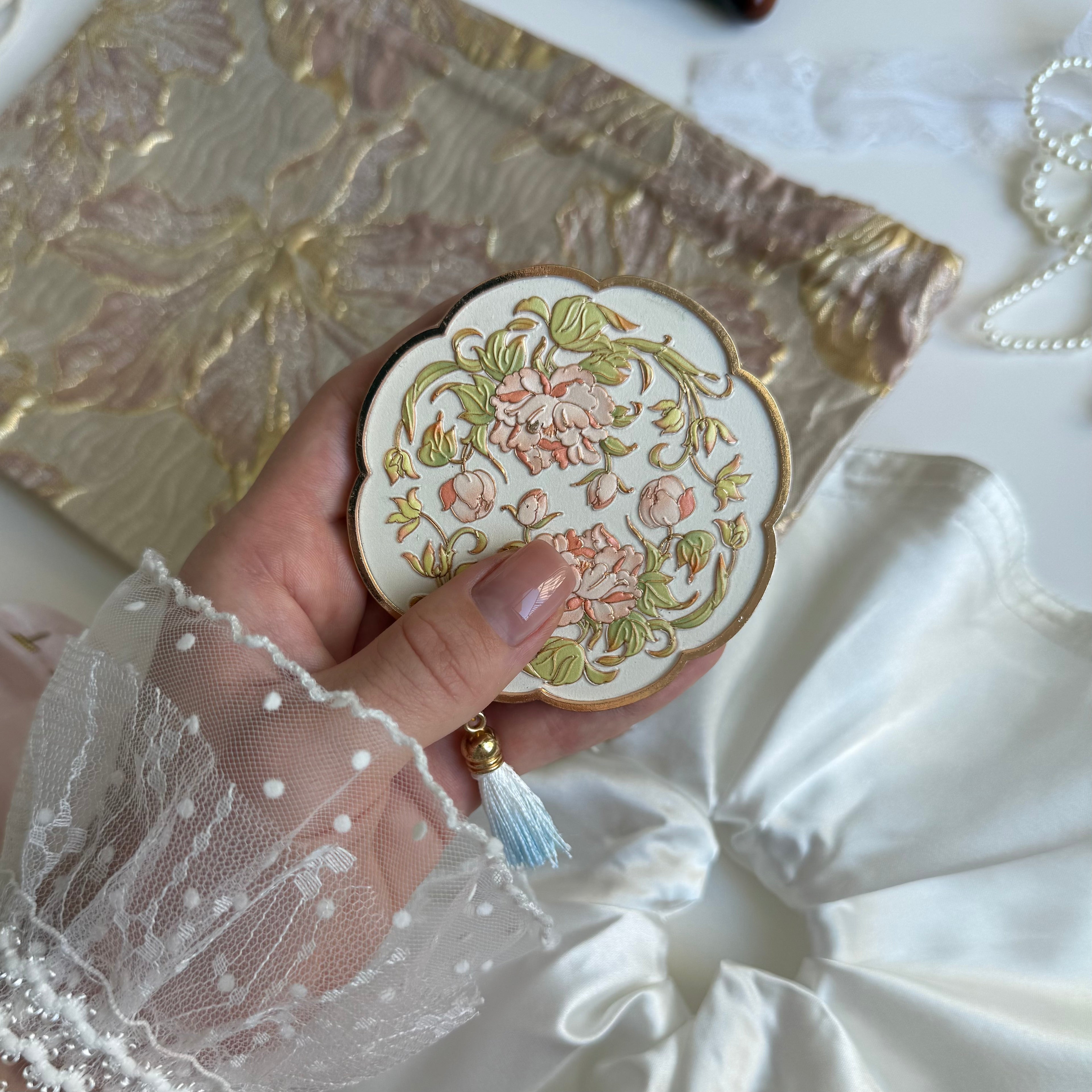 A hand holds a round metal mirror with green and pink flowers. In the background is a jacquard cosmetic bag and a large round satin scrunchie with beautiful pleated folds